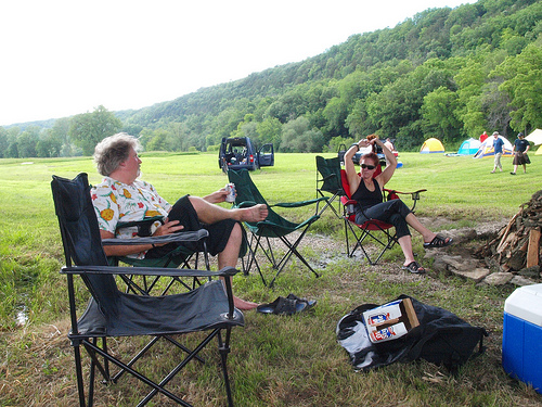  Two people sitting in camp chairs, next to a fire ring in a grass field, with tents and tree covered hills behind